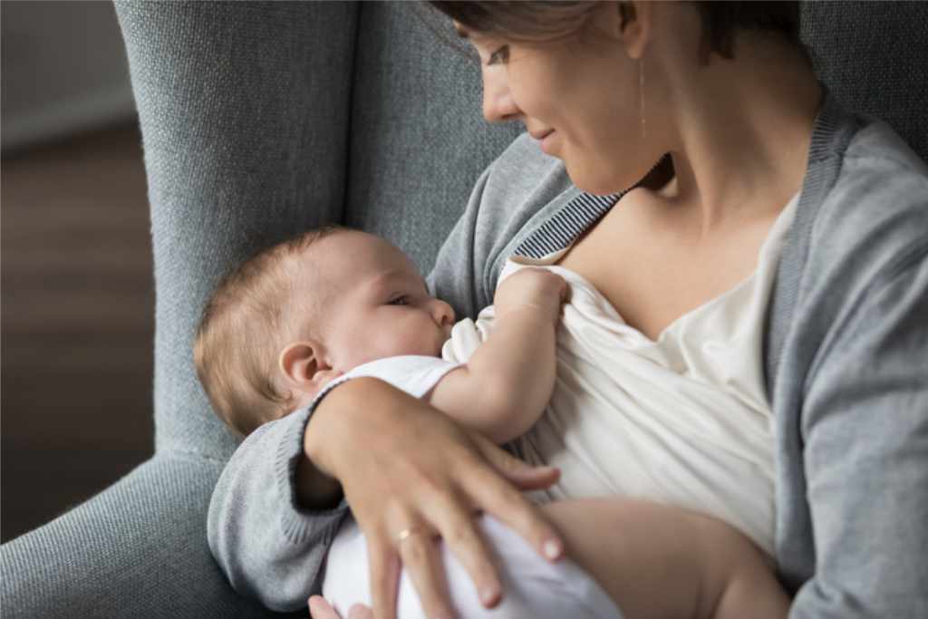 Mom sits in cozy armchair holding baby in tender embrace while breastfeeding.
